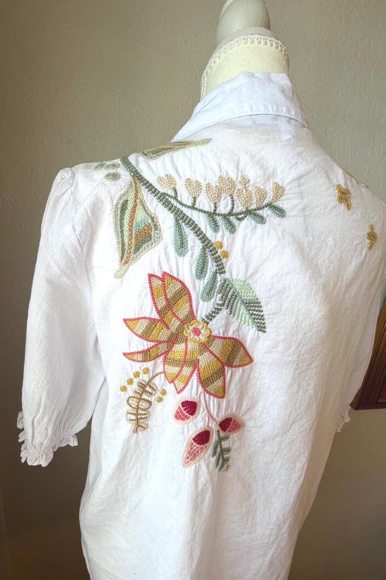 May include: White blouse with floral embroidery on the back. The embroidery features a large flower with yellow, orange, and brown petals, along with green leaves and red accents. The blouse has a collared neckline and short sleeves.