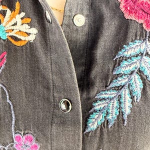 以下が含まれることがあります： カラフルな花の刺繍が施された濃いグレーのデニムジャケットのクローズアップ。刺繍はピンク、オレンジ、ティール、ブルーの花と葉が特徴です。ジャケットにはシルバーのスナップ留めが付いています。