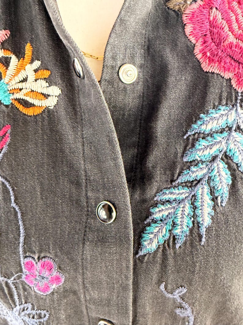 以下が含まれることがあります： カラフルな花の刺繍が施された濃いグレーのデニムジャケットのクローズアップ。刺繍はピンク、オレンジ、ティール、ブルーの花と葉が特徴です。ジャケットにはシルバーのスナップ留めが付いています。