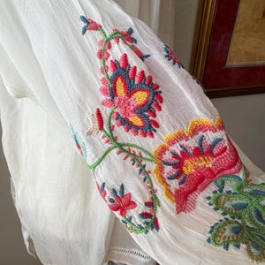 May include: White fabric garment with colorful floral embroidery. The embroidery features red, pink, blue, and green thread, creating a vibrant design. The garment has a flowing, lightweight appearance, suggesting a summer or bohemian style.