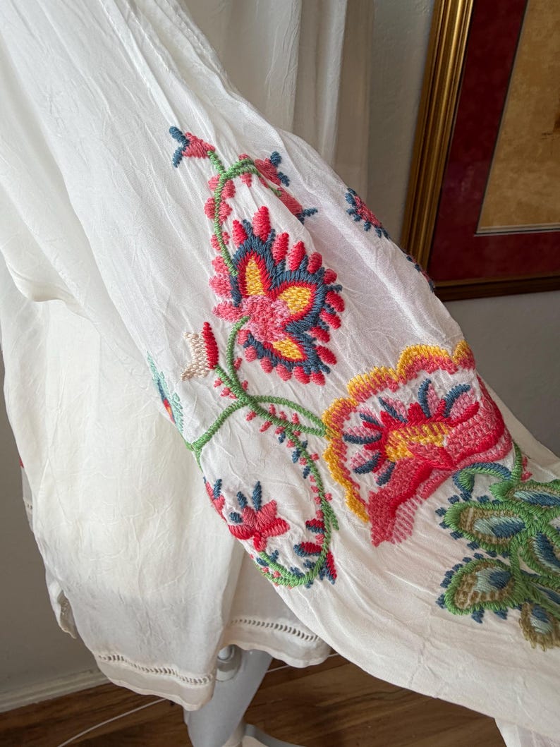 May include: White fabric garment with colorful floral embroidery. The embroidery features red, pink, blue, and green thread, creating a vibrant design. The garment has a flowing, lightweight appearance, suggesting a summer or bohemian style.