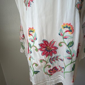 May include: White embroidered tunic with colorful floral designs. The embroidery features red, blue, yellow, and green flowers and leaves. The tunic has a scalloped hemline and is made of a lightweight fabric.