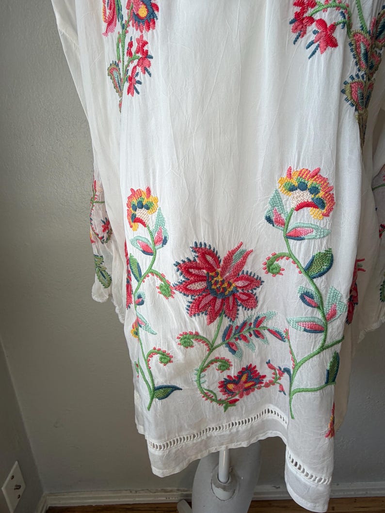 May include: White embroidered tunic with colorful floral designs. The embroidery features red, blue, yellow, and green flowers and leaves. The tunic has a scalloped hemline and is made of a lightweight fabric.