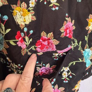 May include: Close-up of a black garment with colorful floral embroidery. The embroidery features yellow, pink, red, blue, and white flowers with green leaves. The garment has a pocket.