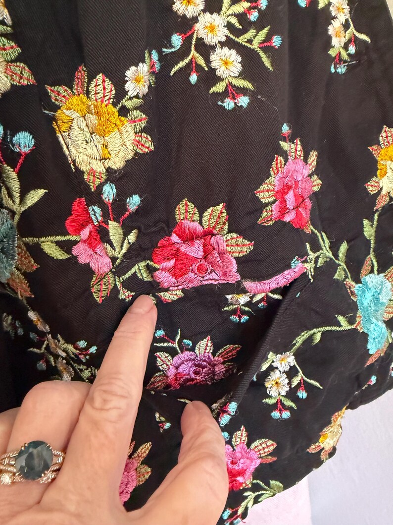May include: Close-up of a black garment with colorful floral embroidery. The embroidery features yellow, pink, red, blue, and white flowers with green leaves. The garment has a pocket.