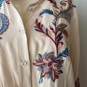 May include: Cream-colored button-down shirt with intricate floral embroidery in shades of red, blue, and brown. The embroidery is detailed and covers the front and collar of the shirt. The shirt has a classic collar and button closure.