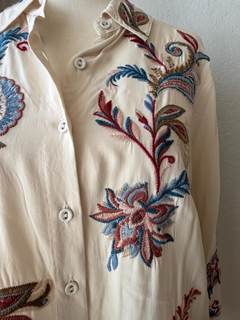 May include: Cream-colored button-down shirt with intricate floral embroidery in shades of red, blue, and brown. The embroidery is detailed and covers the front and collar of the shirt. The shirt has a classic collar and button closure.