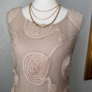 May include: A beige, sleeveless dress with a floral design and a layered gold necklace. The dress has a textured, sheer overlay with raised floral appliques. The necklace features three strands of varying chain styles.