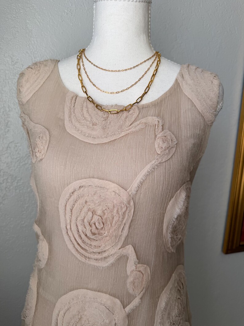 May include: A beige, sleeveless dress with a floral design and a layered gold necklace. The dress has a textured, sheer overlay with raised floral appliques. The necklace features three strands of varying chain styles.