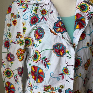 Puede incluir: Chaqueta blanca con bordado floral colorido. La chaqueta presenta un cuello, mangas largas y cierre de botones. El bordado incluye flores y enredaderas rojas, amarillas, azules y verdes, creando un dise&ntilde;o vibrante y art&iacute;stico.