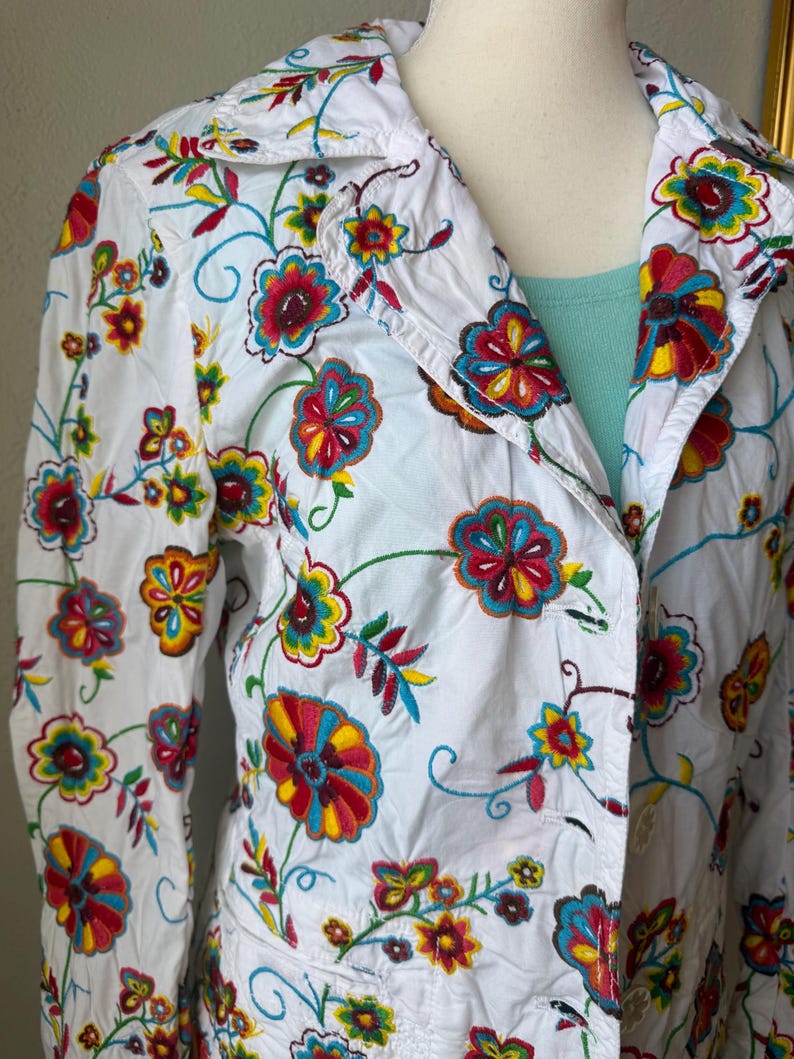 Puede incluir: Chaqueta blanca con bordado floral colorido. La chaqueta presenta un cuello, mangas largas y cierre de botones. El bordado incluye flores y enredaderas rojas, amarillas, azules y verdes, creando un dise&ntilde;o vibrante y art&iacute;stico.