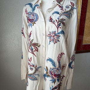 May include: Cream-colored button-down shirt with long sleeves and intricate floral embroidery. The embroidery features blue, red, and green hues, creating a detailed pattern across the shirt's front and sleeves. The shirt is displayed on a mannequin.