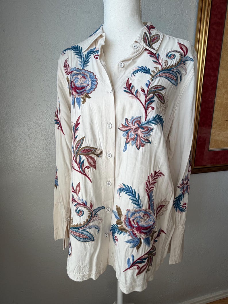 May include: Cream-colored button-down shirt with long sleeves and intricate floral embroidery. The embroidery features blue, red, and green hues, creating a detailed pattern across the shirt's front and sleeves. The shirt is displayed on a mannequin.