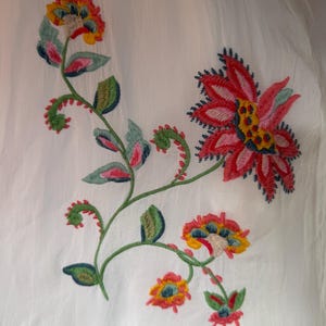 May include: White fabric with colorful floral embroidery. The embroidery features red, pink, yellow, and blue flowers with green leaves and stems. The bottom edge has a decorative, eyelet trim.