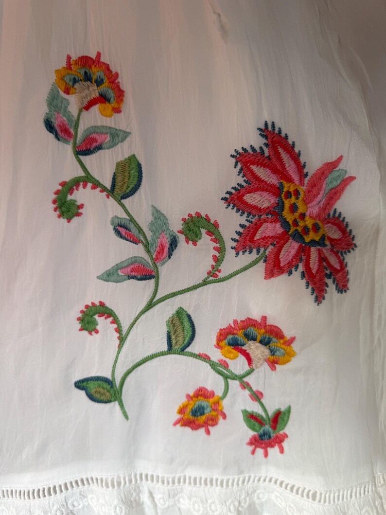 May include: White fabric with colorful floral embroidery. The embroidery features red, pink, yellow, and blue flowers with green leaves and stems. The bottom edge has a decorative, eyelet trim.