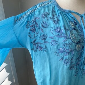 Puede incluir: Una blusa t&uacute;nica azul claro con mangas largas y bordados florales intrincados en tonos azules. La blusa presenta un dise&ntilde;o de cuello anudado y costuras delicadas a lo largo del escote y las mangas. La tela parece ligera y fluida.