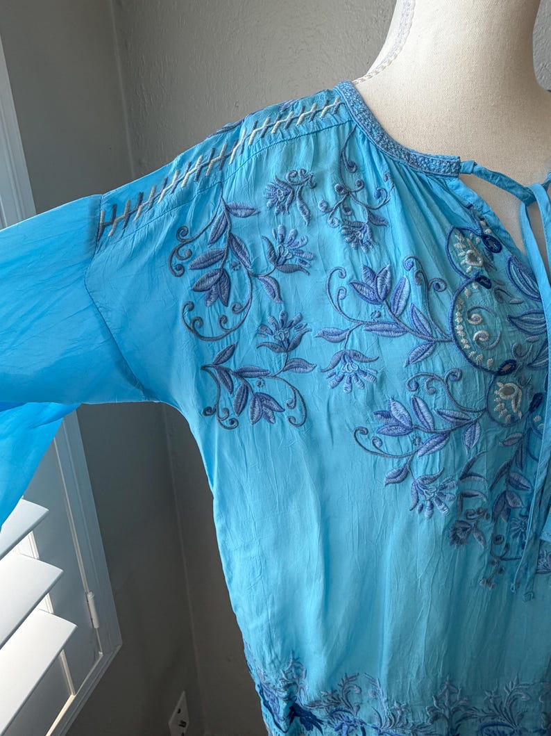 Puede incluir: Una blusa t&uacute;nica azul claro con mangas largas y bordados florales intrincados en tonos azules. La blusa presenta un dise&ntilde;o de cuello anudado y costuras delicadas a lo largo del escote y las mangas. La tela parece ligera y fluida.