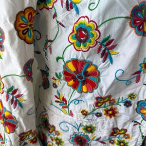 Puede incluir: Tela blanca con bordados florales coloridos. El bordado presenta tonos vibrantes de rojo, amarillo, azul y verde, creando un patr&oacute;n detallado. El dise&ntilde;o incluye flores y hojas de varios tama&ntilde;os.