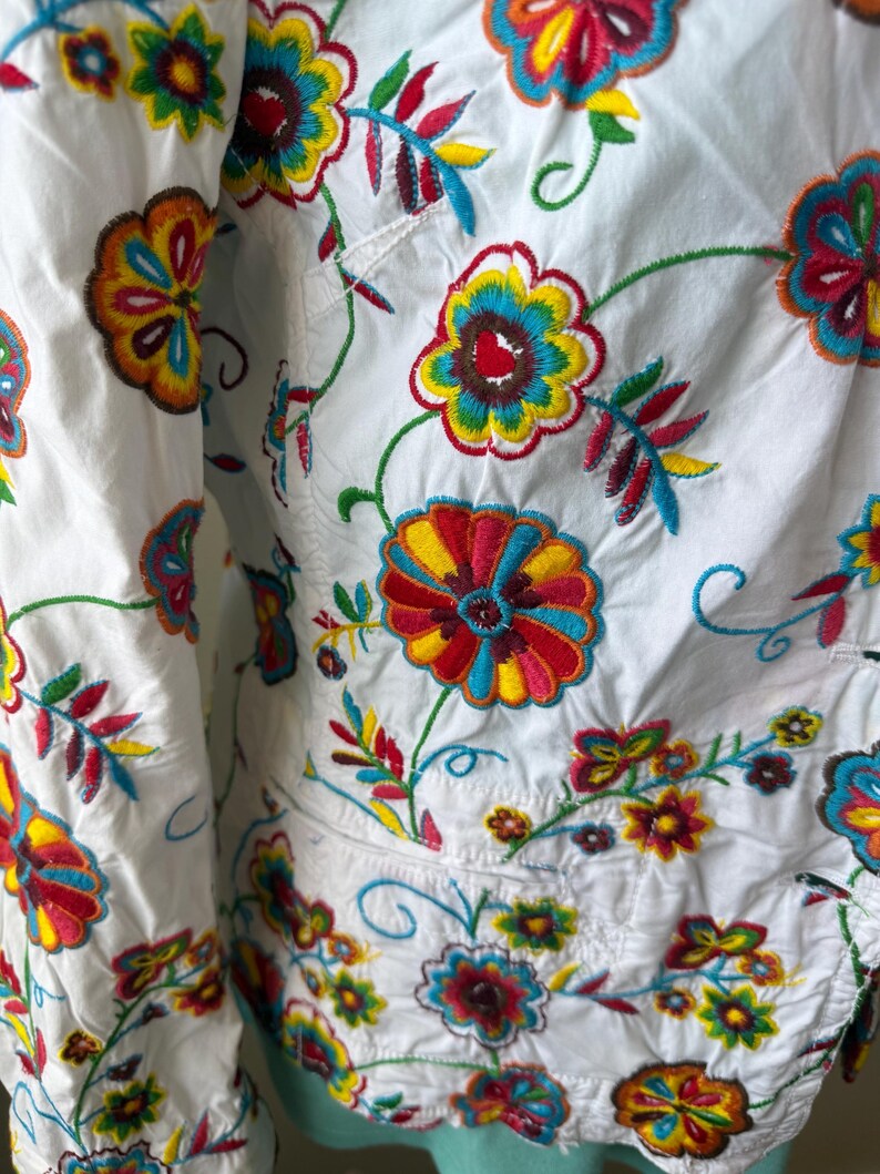 Puede incluir: Tela blanca con bordados florales coloridos. El bordado presenta tonos vibrantes de rojo, amarillo, azul y verde, creando un patr&oacute;n detallado. El dise&ntilde;o incluye flores y hojas de varios tama&ntilde;os.