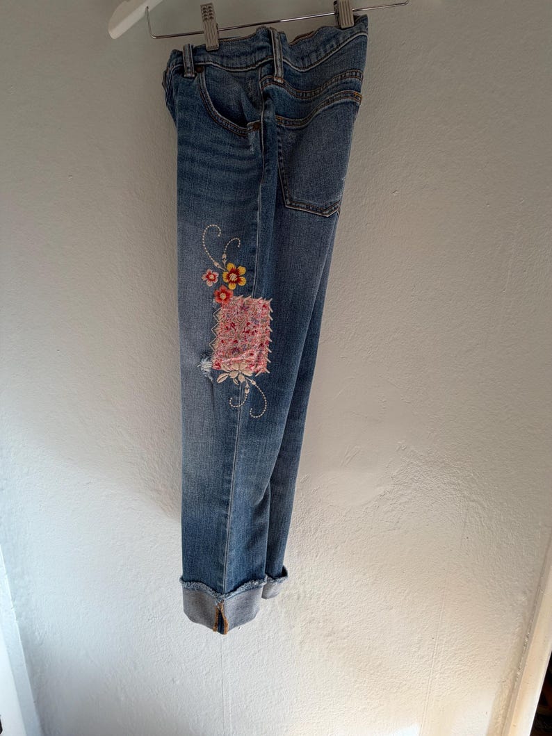 Op de afbeelding: Lichtblauwe spijkerbroek met een bloemenpatch en geborduurde details. De jeans heeft een distressed look met een omgeslagen zoom. De patch heeft roze, gele en rode bloemen.