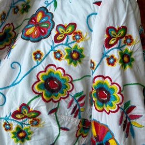 Puede incluir: Camisa blanca con bordados florales coloridos. El bordado presenta flores y enredaderas en tonos rojos, amarillos, azules y verdes. La camisa tiene un bolsillo en el pecho y un cuello.