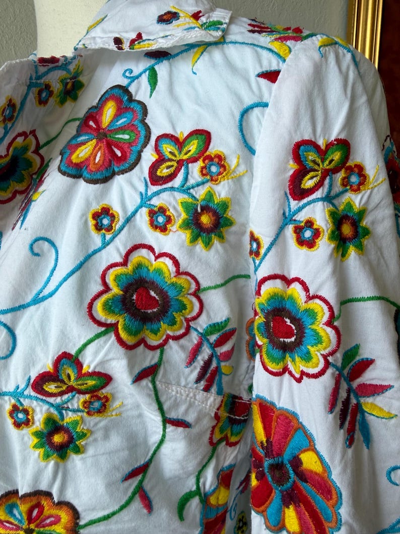 Puede incluir: Camisa blanca con bordados florales coloridos. El bordado presenta flores y enredaderas en tonos rojos, amarillos, azules y verdes. La camisa tiene un bolsillo en el pecho y un cuello.