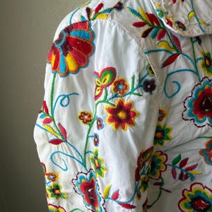 Puede incluir: Camisa blanca con vibrante bordado floral. El dise&ntilde;o colorido presenta flores y hojas rojas, amarillas, azules y verdes. El bordado detallado cubre la parte delantera y las mangas de la camisa.