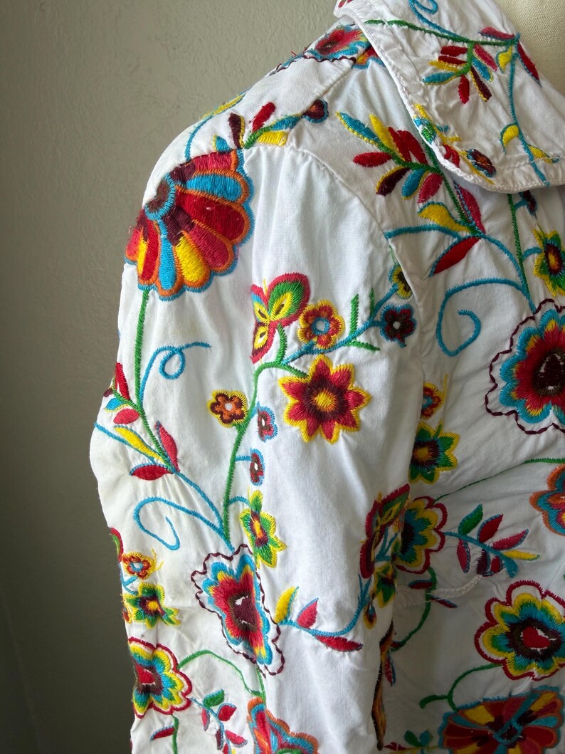 Puede incluir: Camisa blanca con vibrante bordado floral. El dise&ntilde;o colorido presenta flores y hojas rojas, amarillas, azules y verdes. El bordado detallado cubre la parte delantera y las mangas de la camisa.