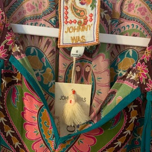 May include: A colorful paisley print top with a V-neckline and turquoise trim. The top features a tag that reads "JOHNNY WAS" and a small tassel. The fabric is a mix of pink, green, brown, and gold.