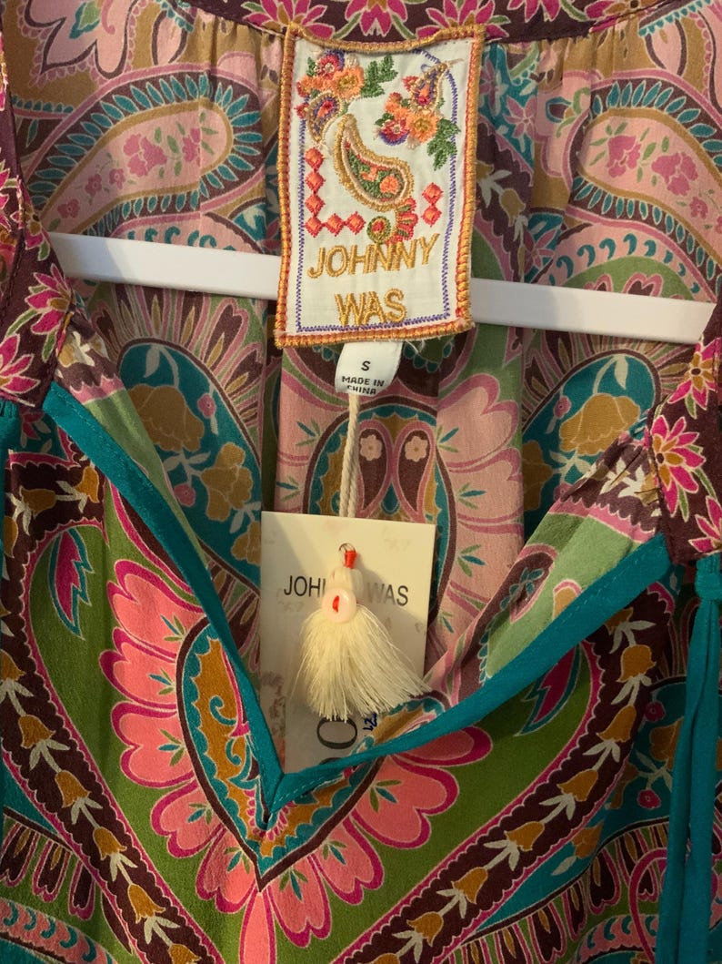 May include: A colorful paisley print top with a V-neckline and turquoise trim. The top features a tag that reads "JOHNNY WAS" and a small tassel. The fabric is a mix of pink, green, brown, and gold.