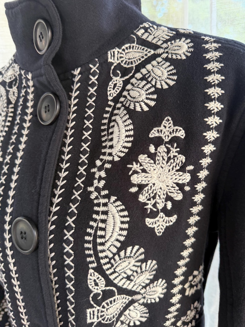 K&ouml;nnte beinhalten: Nahaufnahme einer marineblauen Jacke mit aufwendiger wei&szlig;er Blumenstickerei. Die Jacke hat einen hohen Kragen, drei dunkle Kn&ouml;pfe und dekorative N&auml;hte. Die Stickerei umfasst Blatt- und Blumenmuster.