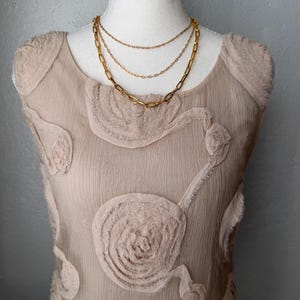 May include: A layered gold-tone necklace with three chains of varying styles, displayed on a beige dress form. The dress is a light beige color with floral-like textured embellishments.