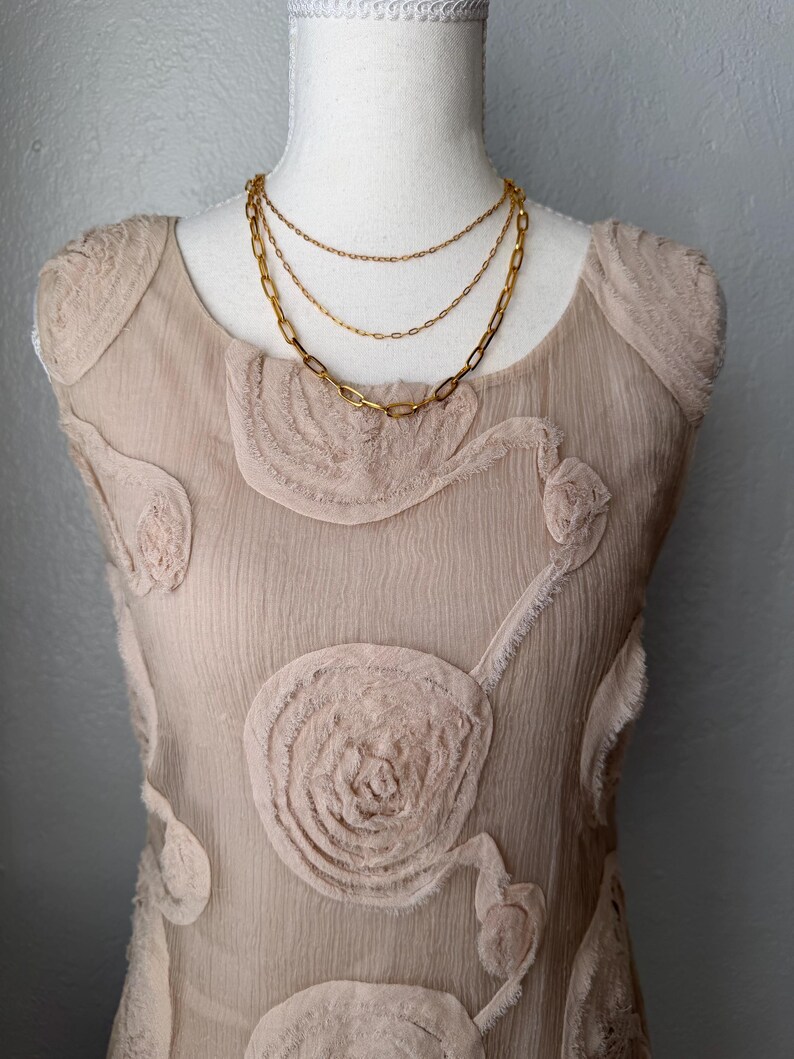May include: A layered gold-tone necklace with three chains of varying styles, displayed on a beige dress form. The dress is a light beige color with floral-like textured embellishments.