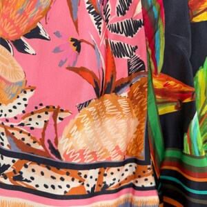 Puede incluir: Primer plano de una tela vibrante con un dise&ntilde;o de frutas y follaje tropical. El patr&oacute;n incluye pi&ntilde;as, hojas y flores en tonos de rosa, naranja, verde y negro. La tela tiene un borde a rayas con varios colores.