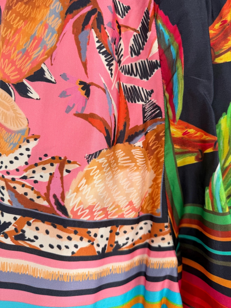 Puede incluir: Primer plano de una tela vibrante con un dise&ntilde;o de frutas y follaje tropical. El patr&oacute;n incluye pi&ntilde;as, hojas y flores en tonos de rosa, naranja, verde y negro. La tela tiene un borde a rayas con varios colores.