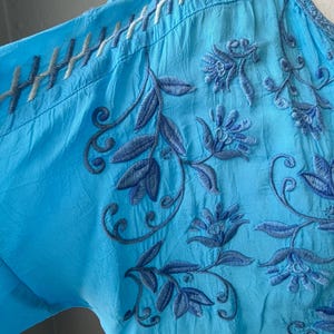 Puede incluir: Una blusa azul turquesa con intrincados bordados florales en tonos azules. La blusa presenta un cuello en V y costuras detalladas a lo largo de los hombros y el escote. La tela parece ligera y ligeramente texturizada.