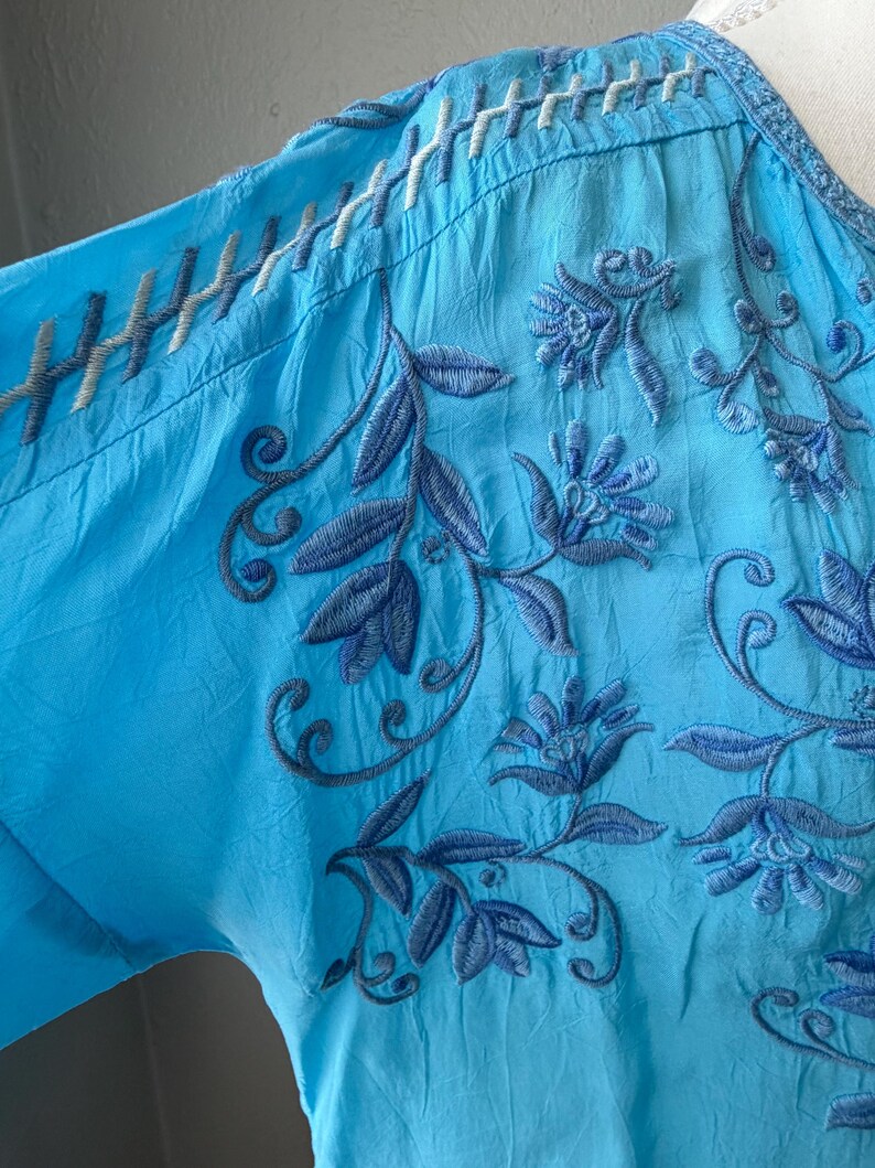 Puede incluir: Una blusa azul turquesa con intrincados bordados florales en tonos azules. La blusa presenta un cuello en V y costuras detalladas a lo largo de los hombros y el escote. La tela parece ligera y ligeramente texturizada.