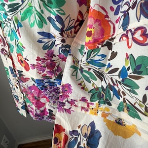 May include: Close-up of a cream-colored fabric with a vibrant floral pattern. The design features large, colorful flowers in shades of pink, orange, blue, purple, and green, set against a neutral background. The fabric appears to be a lightweight material.