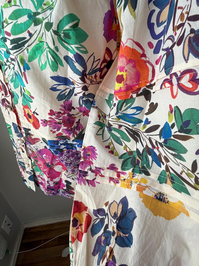 May include: Close-up of a cream-colored fabric with a vibrant floral pattern. The design features large, colorful flowers in shades of pink, orange, blue, purple, and green, set against a neutral background. The fabric appears to be a lightweight material.