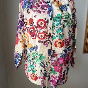 May include: A cream-colored button-down shirt with a vibrant floral pattern. The long-sleeved blouse features a mix of red, purple, blue, and yellow flowers with green leaves. The shirt has a mandarin collar and is displayed on a mannequin.