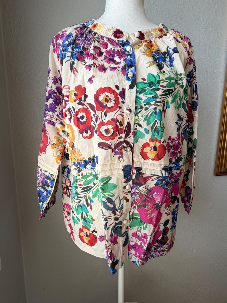 May include: A cream-colored button-down shirt with a vibrant floral pattern. The long-sleeved blouse features a mix of red, purple, blue, and yellow flowers with green leaves. The shirt has a mandarin collar and is displayed on a mannequin.