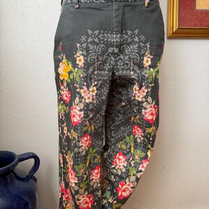 Puede incluir: Pantalones gris carb&oacute;n oscuro con un estampado floral de rosas rojas, rosas y amarillas, hojas verdes y un patr&oacute;n sim&eacute;trico gris de estilo damasco. Los pantalones incluyen una cintura con trabillas, cierre de bot&oacute;n y bolsillos laterales.
