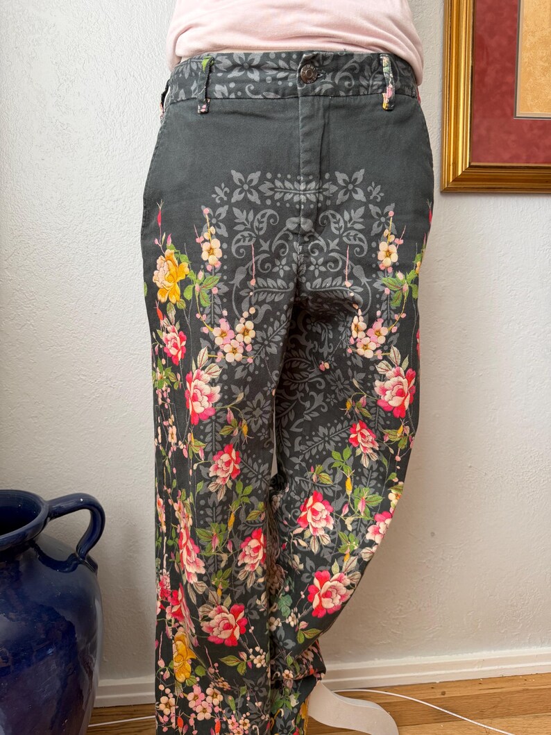 Puede incluir: Pantalones gris carb&oacute;n oscuro con un estampado floral de rosas rojas, rosas y amarillas, hojas verdes y un patr&oacute;n sim&eacute;trico gris de estilo damasco. Los pantalones incluyen una cintura con trabillas, cierre de bot&oacute;n y bolsillos laterales.