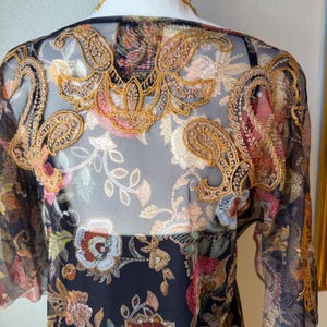 May include: A sheer black top with intricate floral embroidery in shades of gold, red, pink, and blue. The top features short sleeves and a detailed embroidered design across the shoulders and back, showcasing a bohemian style.