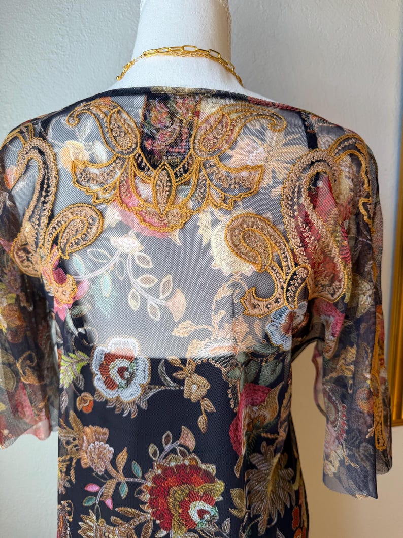 May include: A sheer black top with intricate floral embroidery in shades of gold, red, pink, and blue. The top features short sleeves and a detailed embroidered design across the shoulders and back, showcasing a bohemian style.