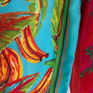 Puede incluir: Un textil vibrante con un estampado de pl&aacute;tanos coloridos sobre un fondo azul claro. El dise&ntilde;o incluye pl&aacute;tanos rojos, amarillos y naranjas con hojas verdes. Un borde a rayas en verde, naranja y marr&oacute;n enmarca el dise&ntilde;o. A la derecha se ve una tela roja con estampado de chiles.