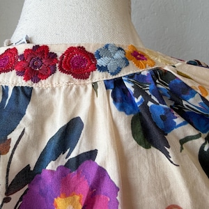 May include: Close-up of a cream-colored blouse with colorful floral embroidery along the neckline. The blouse features large, vibrant floral patterns in shades of purple, blue, and red. The fabric appears to be a soft, lightweight material.