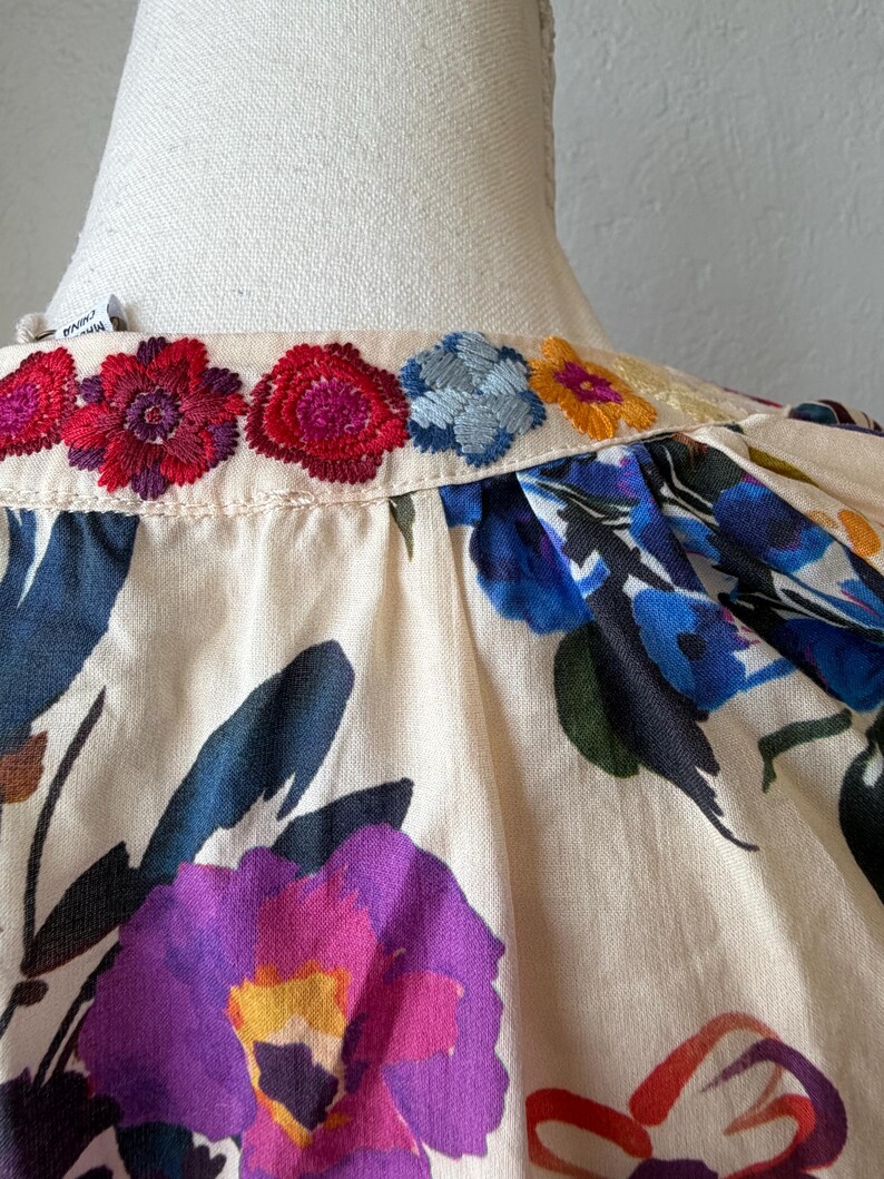 May include: Close-up of a cream-colored blouse with colorful floral embroidery along the neckline. The blouse features large, vibrant floral patterns in shades of purple, blue, and red. The fabric appears to be a soft, lightweight material.