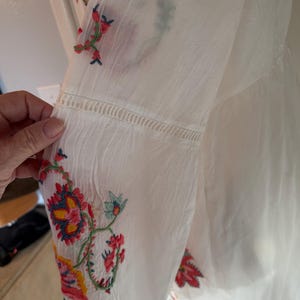 May include: White long-sleeved blouse with colorful floral embroidery on the sleeve. The embroidery features red, yellow, blue, and green thread. The sleeve has a decorative stitched detail near the elbow.