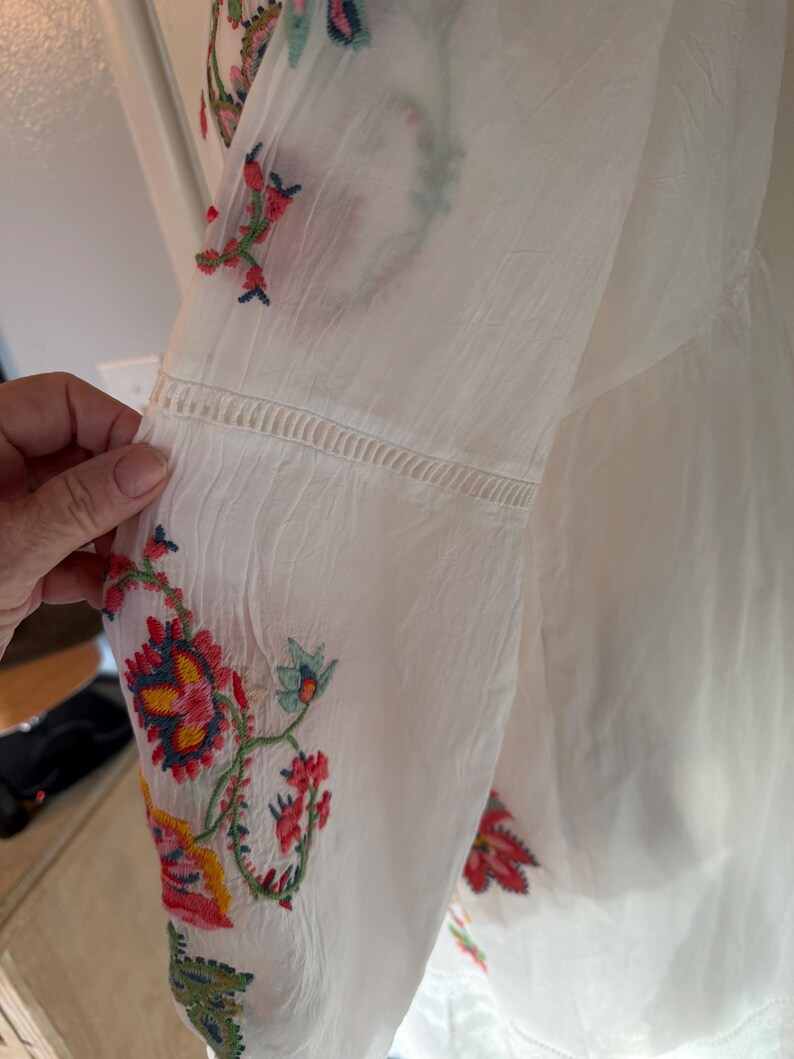 May include: White long-sleeved blouse with colorful floral embroidery on the sleeve. The embroidery features red, yellow, blue, and green thread. The sleeve has a decorative stitched detail near the elbow.